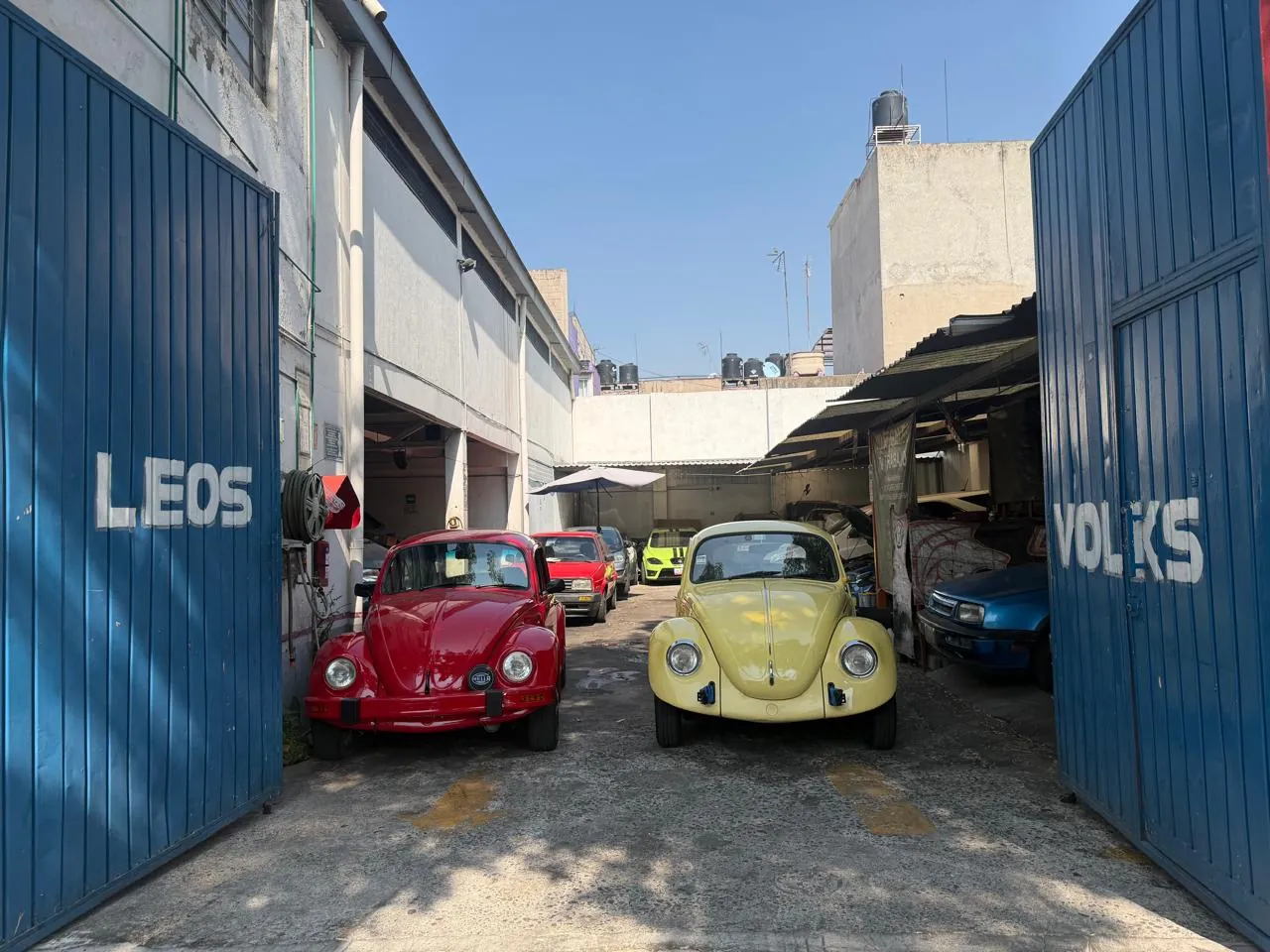 Entrada del taller Leos Volks con dos vochos clásicos, rojo y amarillo, estacionados frente al portón